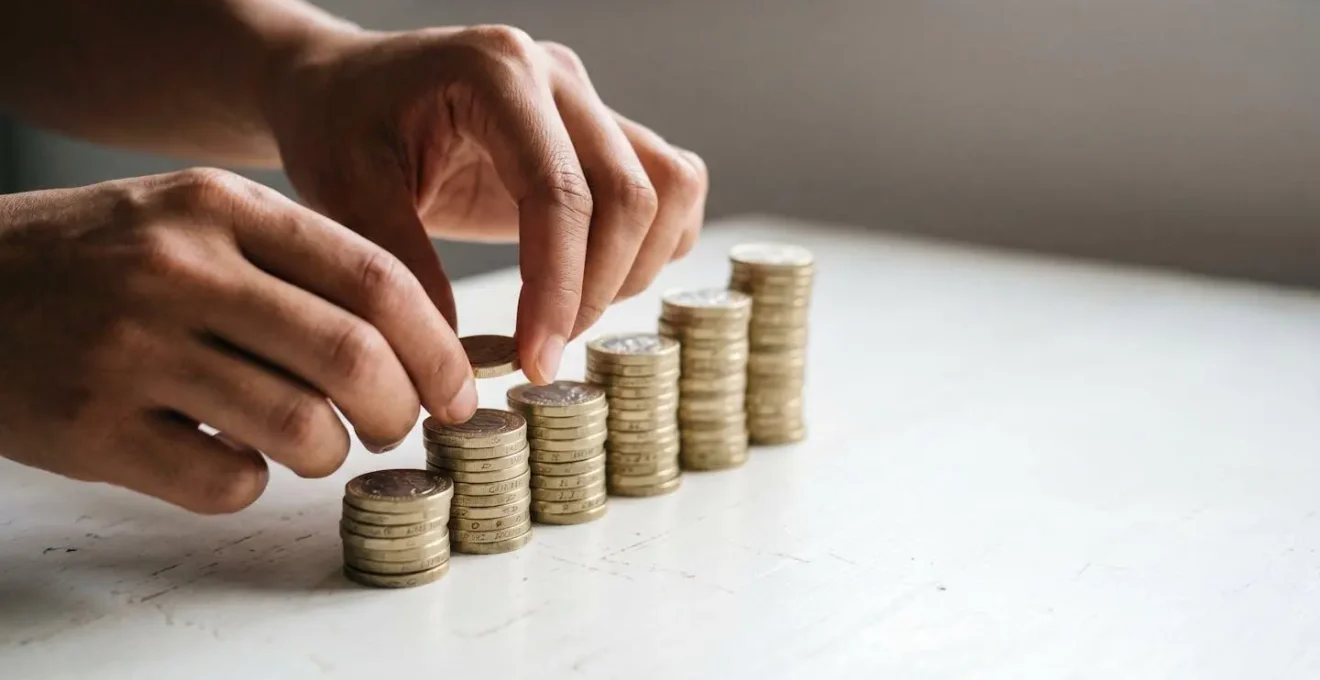 Hands carefully stacking British pound coins in ascending piles against a minimal background, symbolizing building emergency savings during economic uncertainty