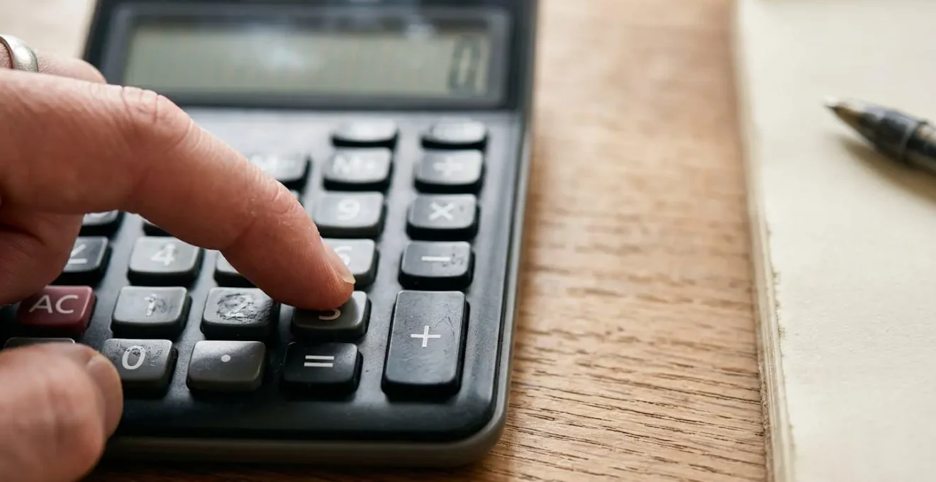 Close-up of hands using a calculator alongside organized financial documents on a wooden desk, representing the calculation of personalized emergency fund targets